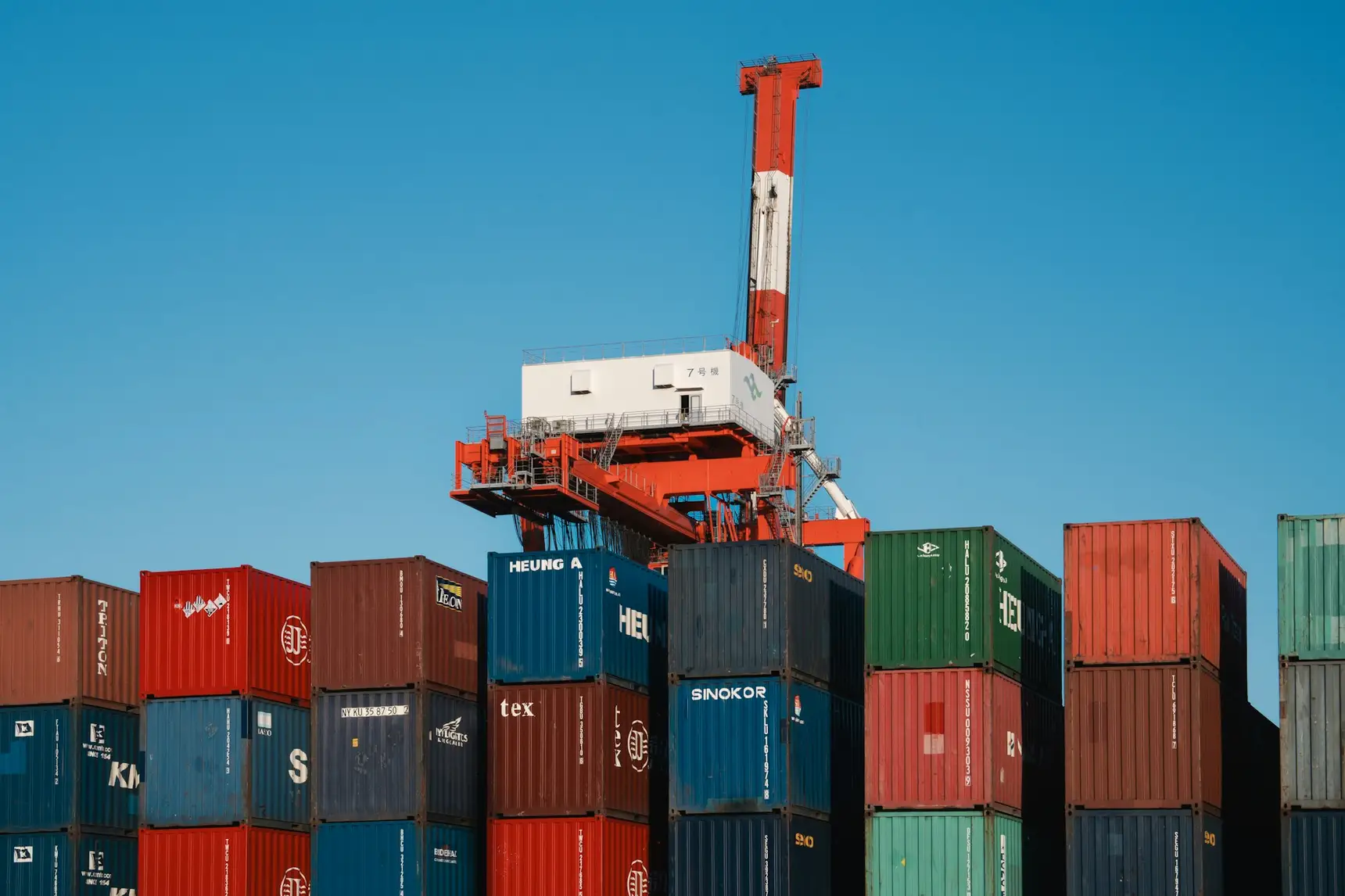 Shipping containers stacked several high at a port under a gantry crane, marked with Asian shipping lines including Heung-A, SINOKOR and KMTC