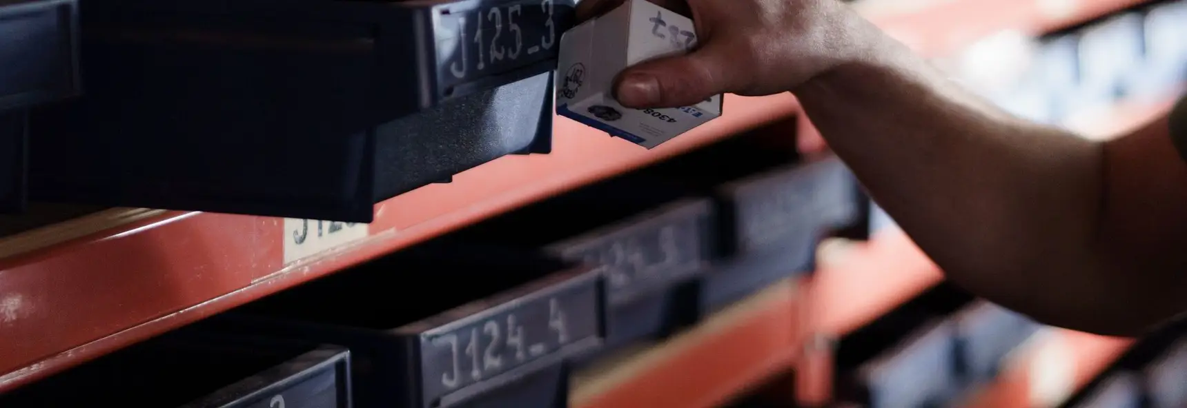 A hand lifting a small boxed component out of bin J125_3 on warehouse racking, among rows of identically labelled bins.